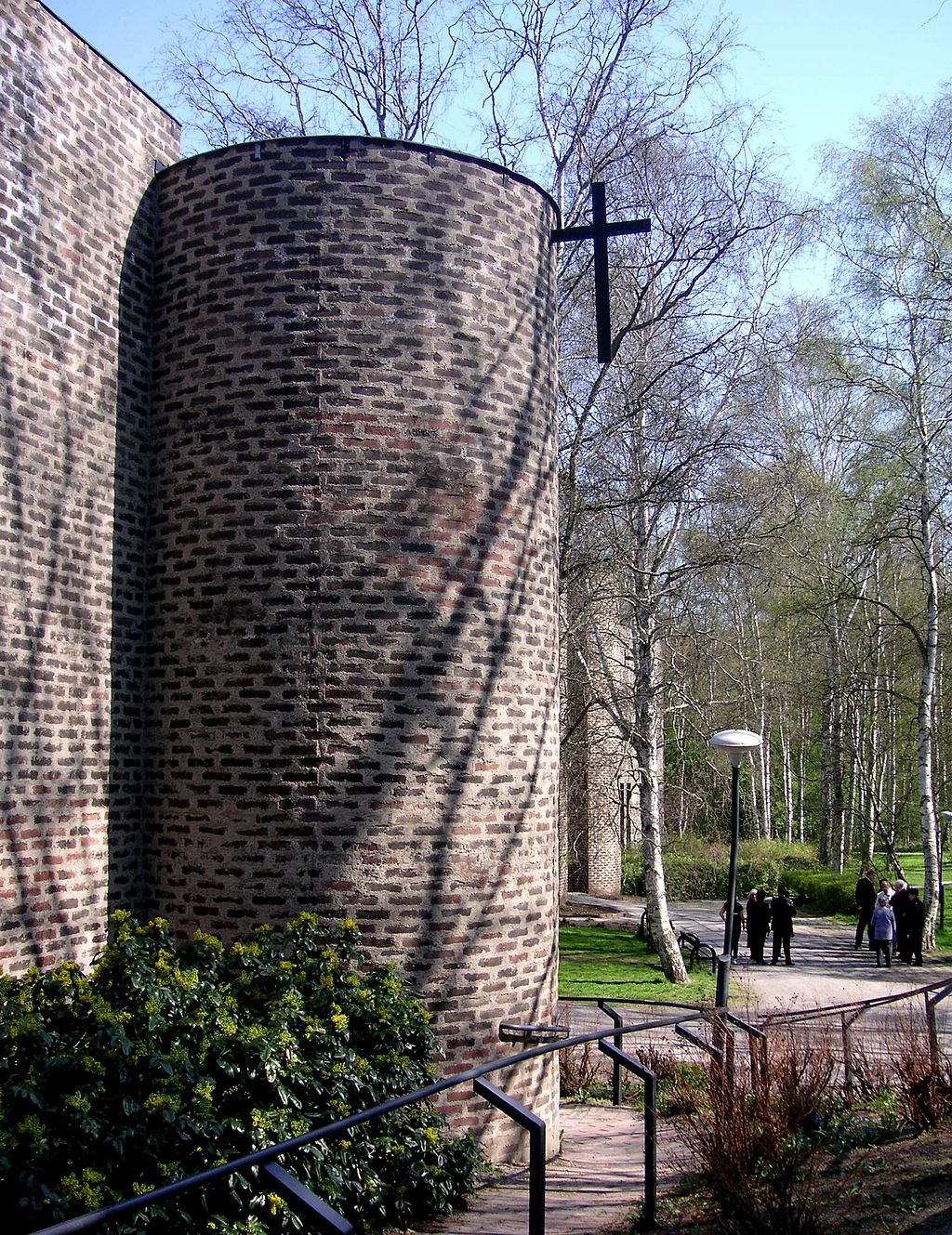 Ett fönsterlöst cylindriskt torn i brunt tegel ungefär 7 meter högt och ett kors som är fäst i tornets sida med ena armen. Träden i närheten kastar skuggor över fasaden.