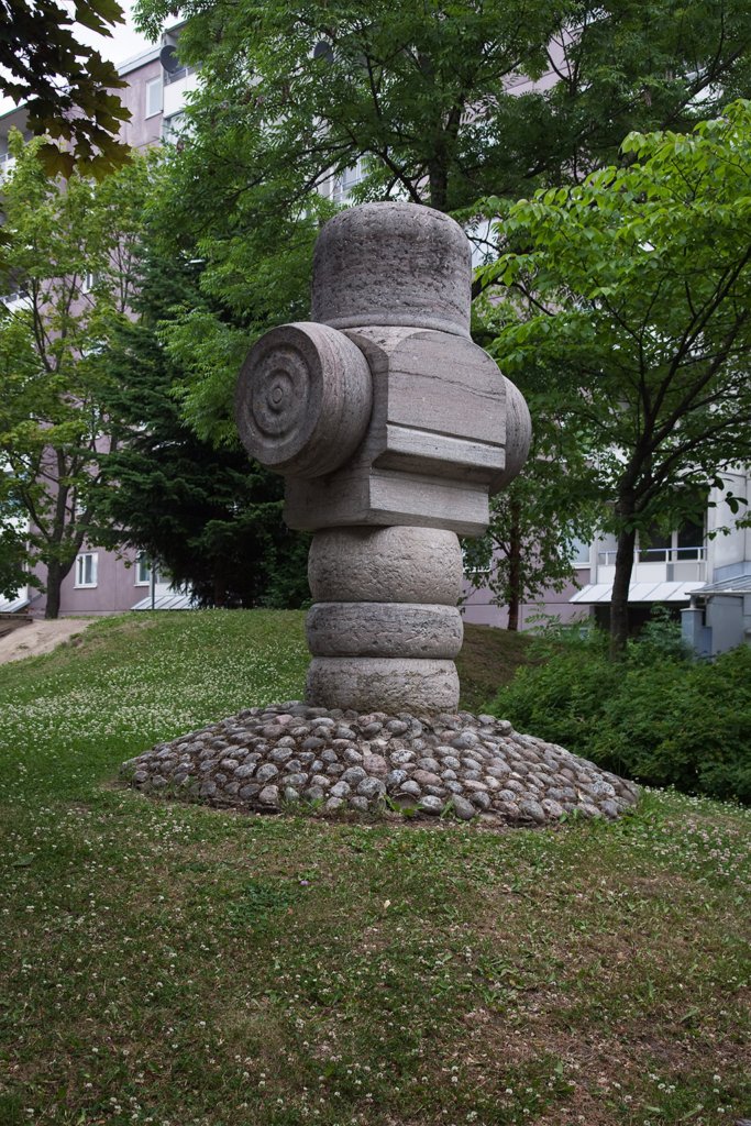 Skulptur av sten med fyrkantiga och runda former som står på gräs. Träd och hus i bakgrunden.
