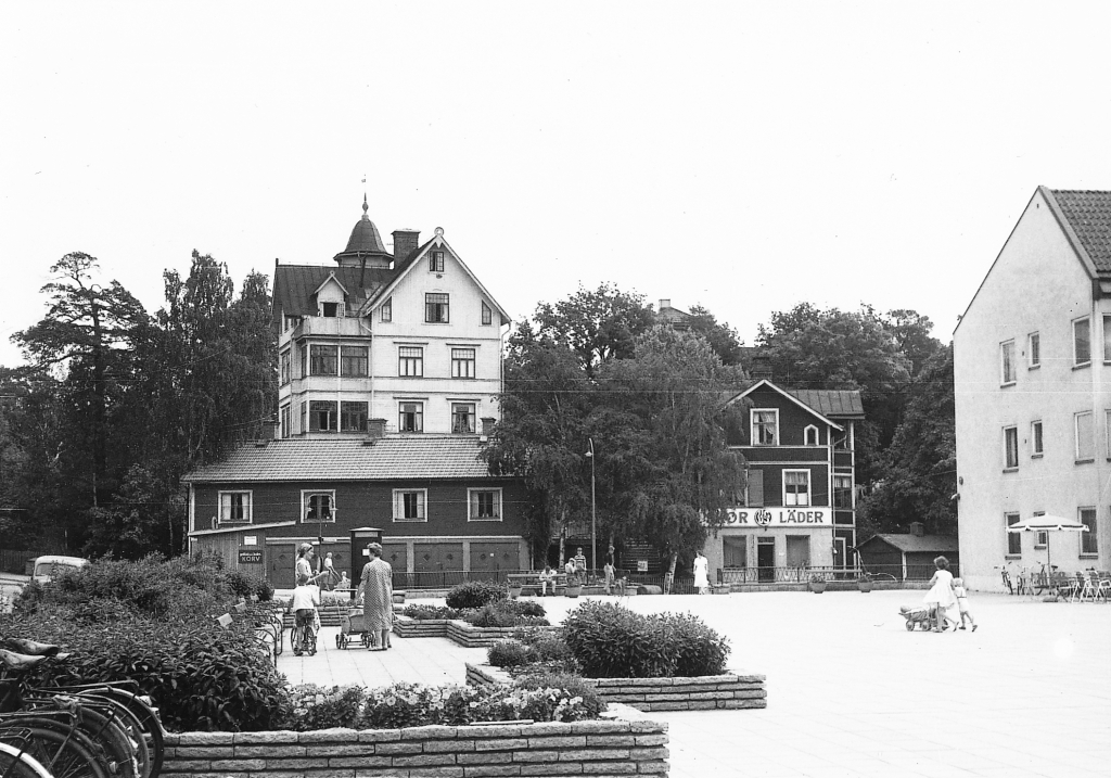 Barn och kvinnor på ett torg med planteringar och bänkar. I bakgrundens syns tre trähus, ett ligger på en kulle och har fyra våningar och ett torn. De andra ligger mot gatan i torgets fond. Det ena huset har fyra garageportar mot vägen och det andra har en skylt där det står "skor" och "läder".