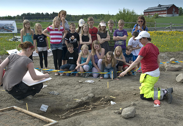 En grupp med barn står och tittar på en utgrävning. Två personer visar och berättar.