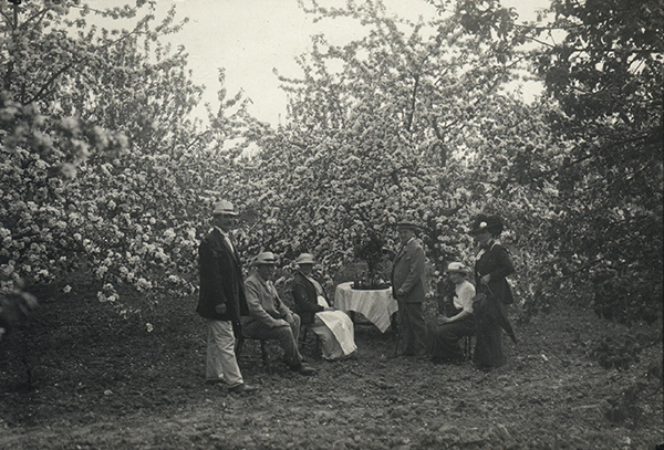 Historisk bild på en grupp människor som sitter i en trädgård. De är finklädda.