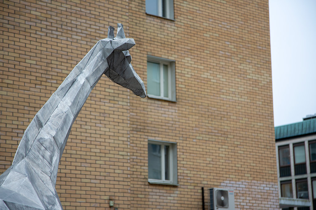 Skulptur av giraff i aluminium, där bara halsen och huvudet är synligt. Gul tegelvägg bakom.