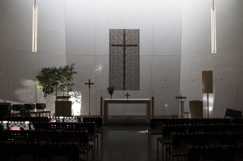 Fotot visar kyrkans enkla altare mot vita väggar och en bonad med ett kors.