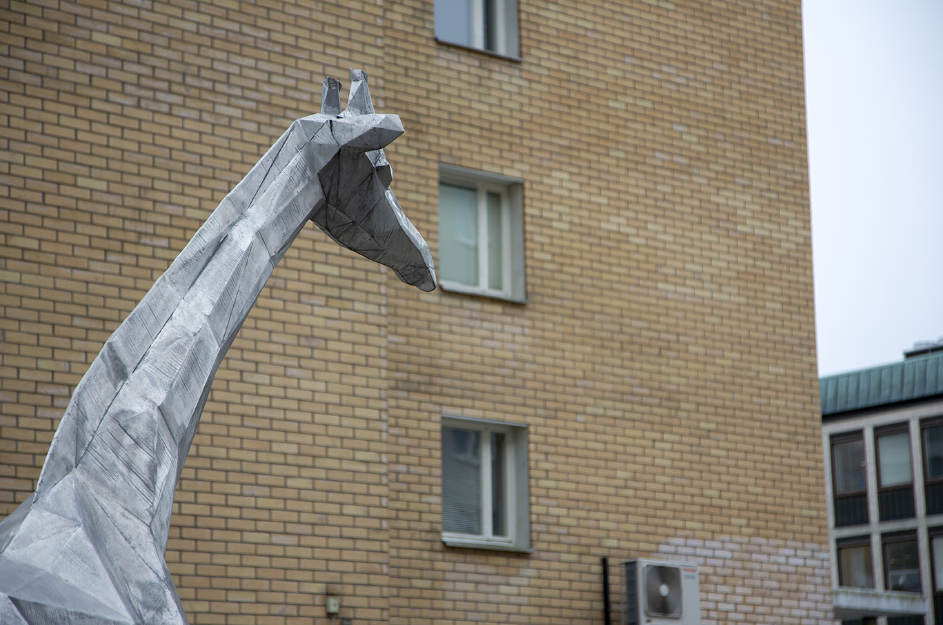 En skulptur i aluminium som föreställer en giraff som tittar över ett torg.
