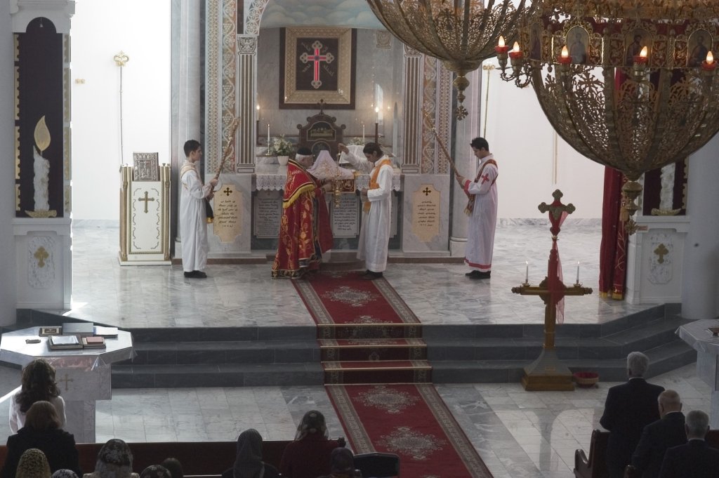 Prästen, i röd klädnad står på ett podium längst fram i kyrkan tillsammans med tre vitklädda korgossar.