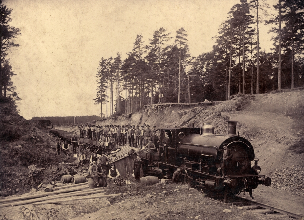 Ett ånglok står på spåret som skär genom en tallbevuxen ås. På tågets lastvagnar och framför loket står ett fyrtiotal arbetare uppställda för fotografering. Bildnummer: 38