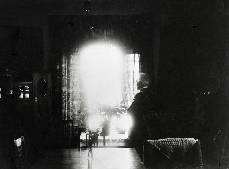 En mansgestalt skymtar i ett rum, starkt motljus från ett fönster. Bilden är ett äldre fotografi, mörk och mystisk.