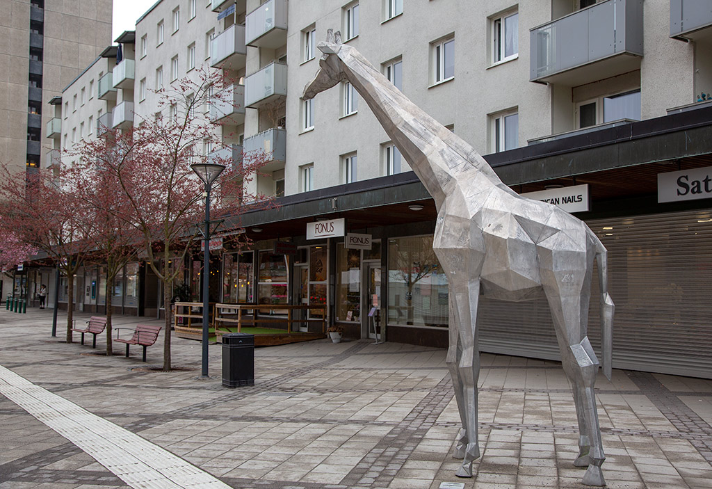 Skulptur av giraff i aluminium på ett torg med affärer bakom.