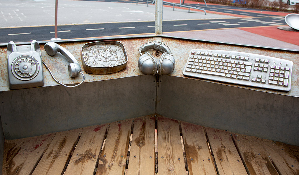 Telefon, hörlurar och tangentbord i metall på en metallbänk ovanför ett trägolv. Utomhus.