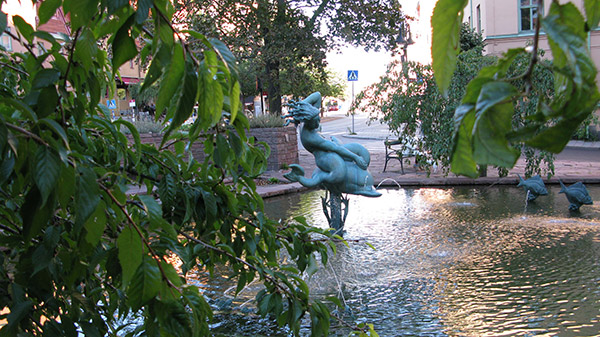 Fontän med skulptur av en flicka som rider på en delfin, även skulpturer av två fiskar i dammen. Grönska och bladverk runtomkring.