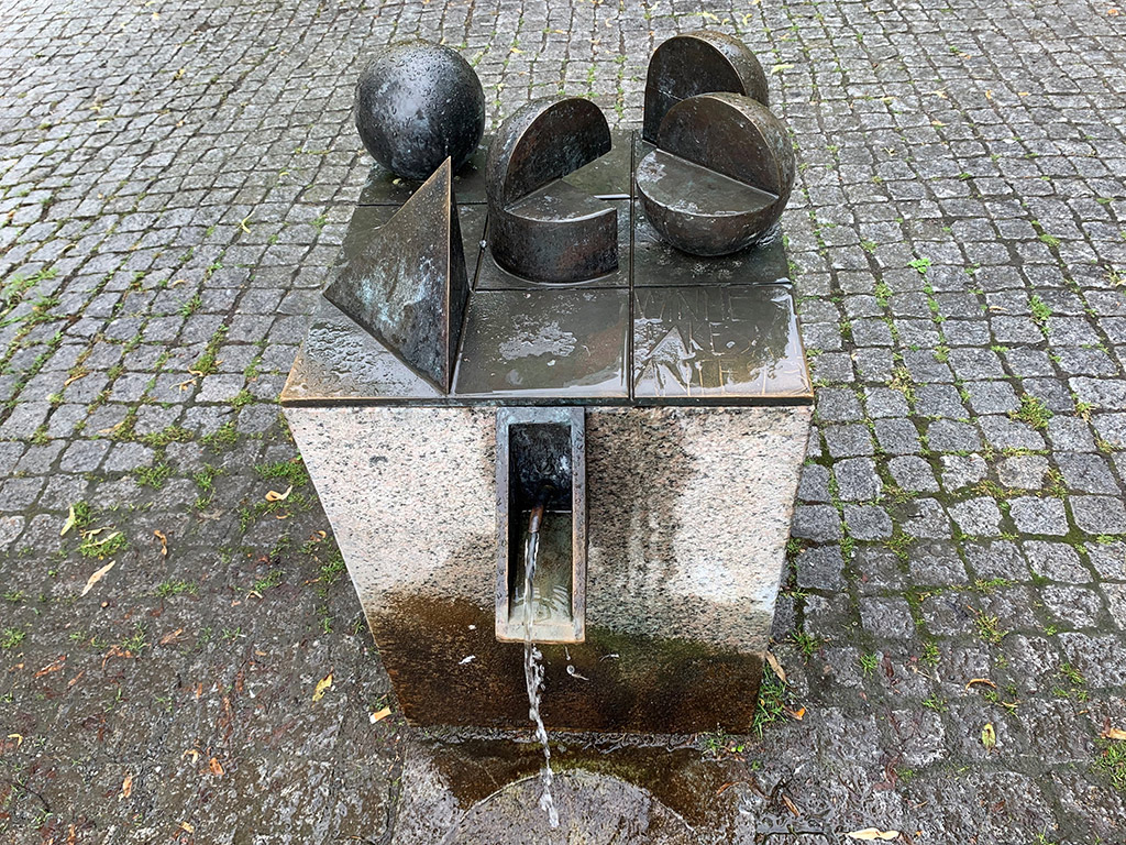 Fem geometriska former, klot, halvklot av brons på en bronsplatta med sex fyrkantiga fält. Under sprutar vatten ner på marken i en rund stenskål.