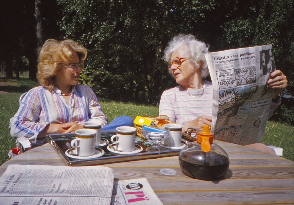 Två kvinnor sitter och fikar i det gröna och bläddrar i en dagstidning.
