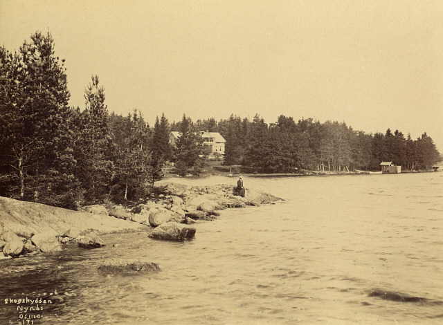 ett svartvitt foto. Ett ensamt hus ligger vid vattnet omgivet av skog. På en klippa står en kvinna.