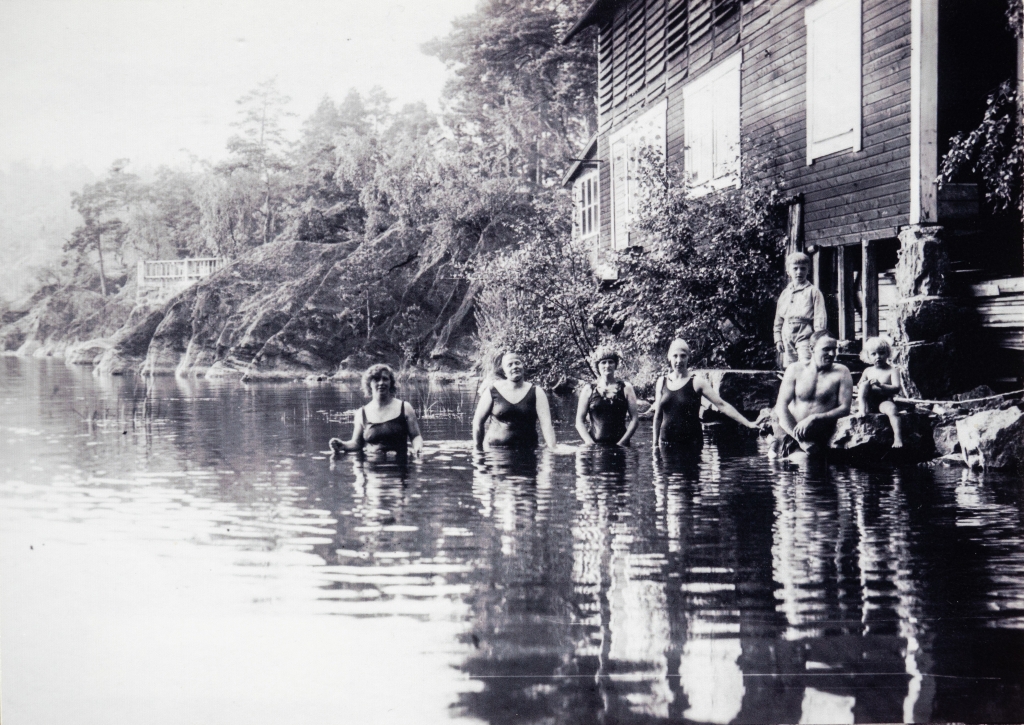 Fyra kvinnor och en man i badkälder står i vattnet och två barn sitter på stranden.