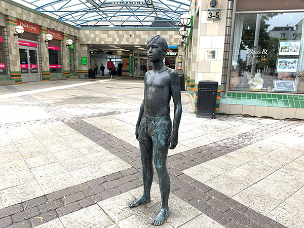 Skulptur i brons av en pojke som står utanför en butik.