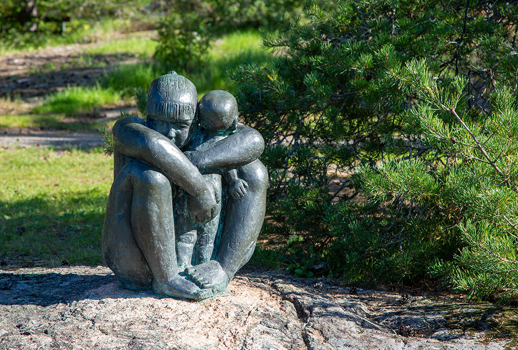 En skulptur av en kvinna med barn som sitter på en sten. En tall bredvid och gräs bakom.