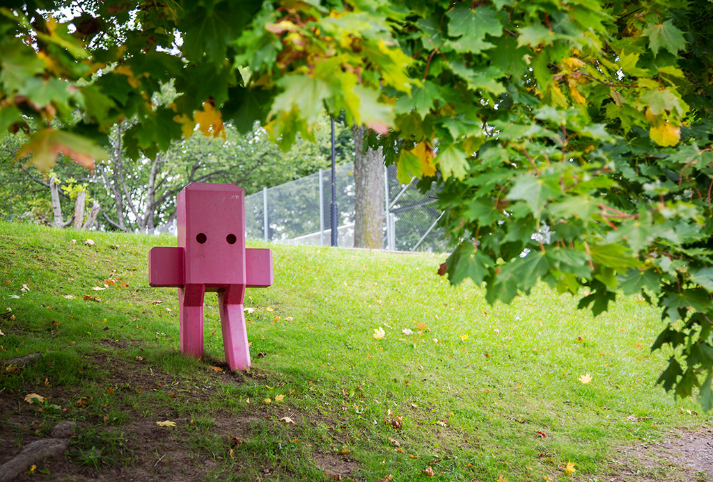 En rosa skulptur med två runda ögon, ben och med armar som pekar rakt ut. Den står i en slänt i skuggan av ett lövträd.