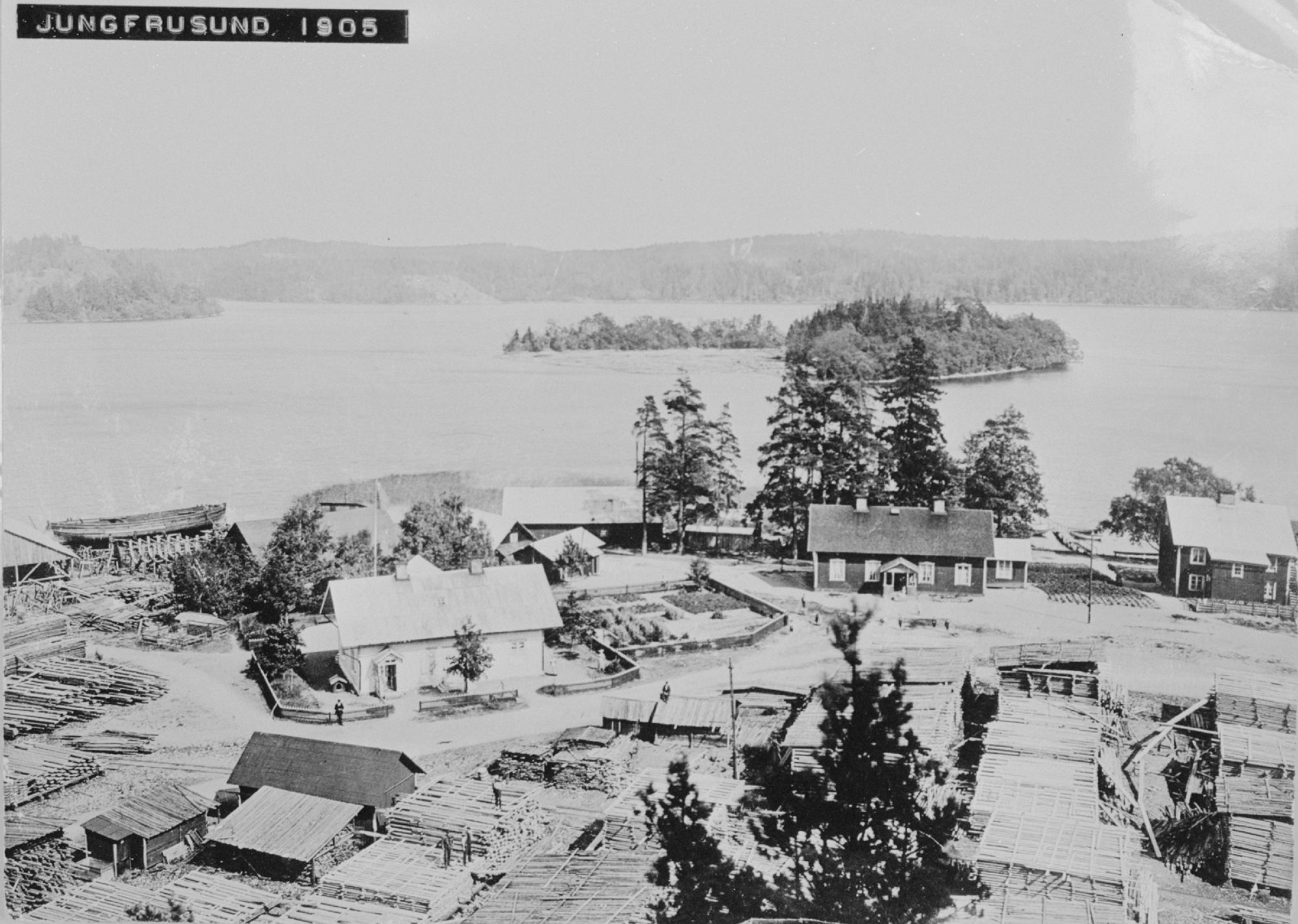 Ett svartvitt foto av några hus vid vattnet omgivna av högar av trä och virke.