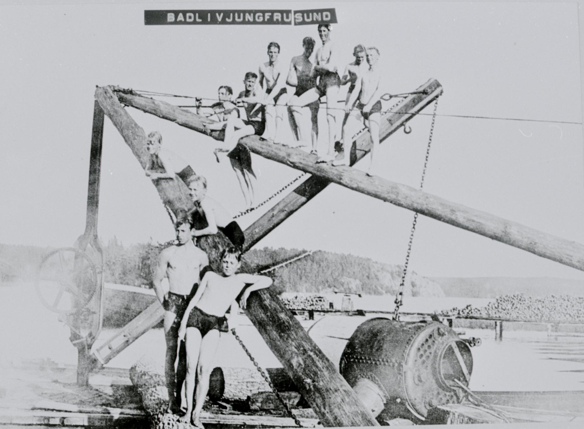 Svartvitt foto av elva pojkar i badbyxor som klättrar på en kran intill vattnet.