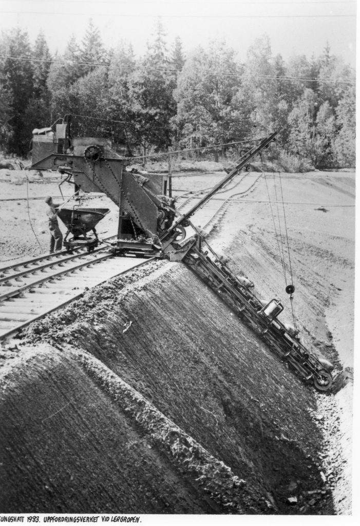 Fotot visar en maskin som går på en räls längst ett långt dike där maskinen sträcker sig ner för att ta upp lera och lasta på en vagn på en smalare parallell räls.