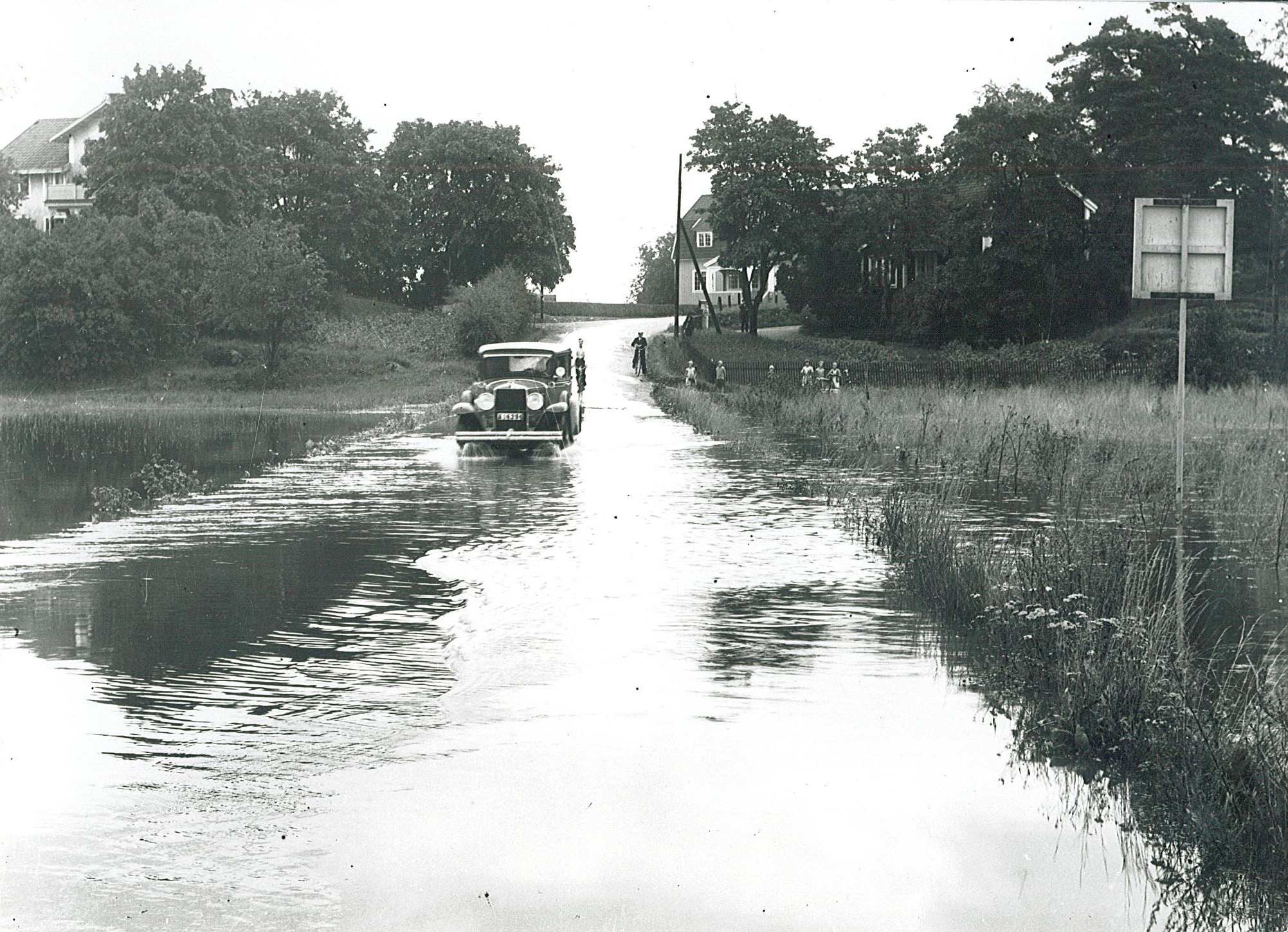 En ålderdomlig bil kör genom en helt översvämmad väg över en äng.