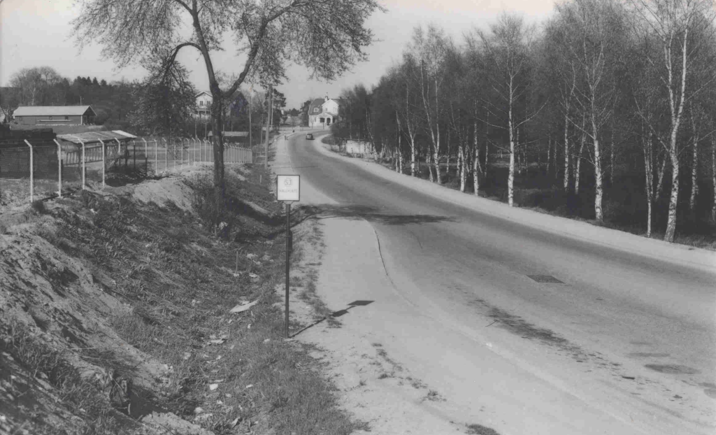 Ett svartvitt foto av en landsväg som leder upp för en backe till några hus. Mitt i bilden står en skylt.