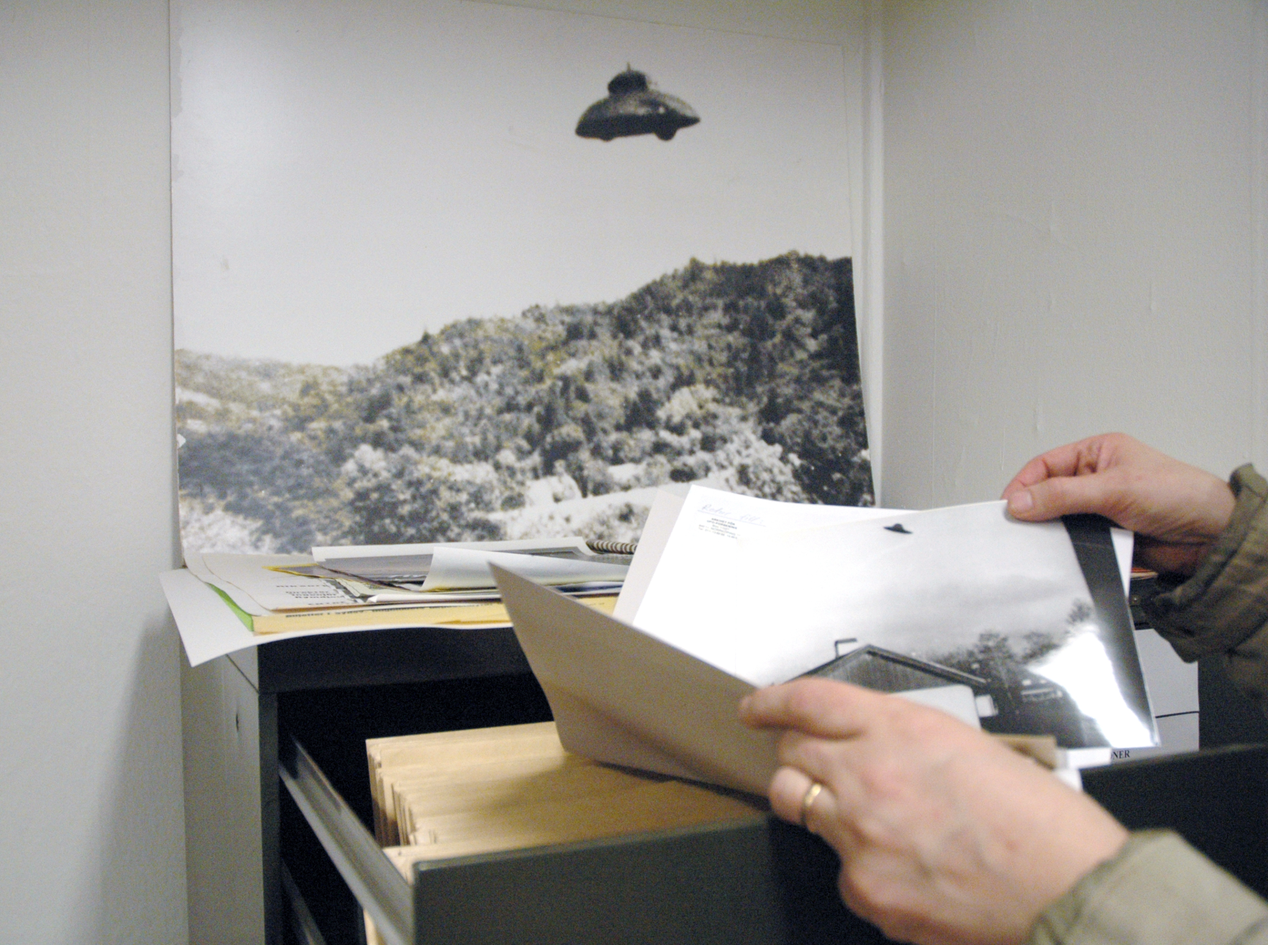 En person står i ett arkiv och bläddrar bland bilder. På väggen sitter ett fotografi som ser ut att förställa ett UFO.