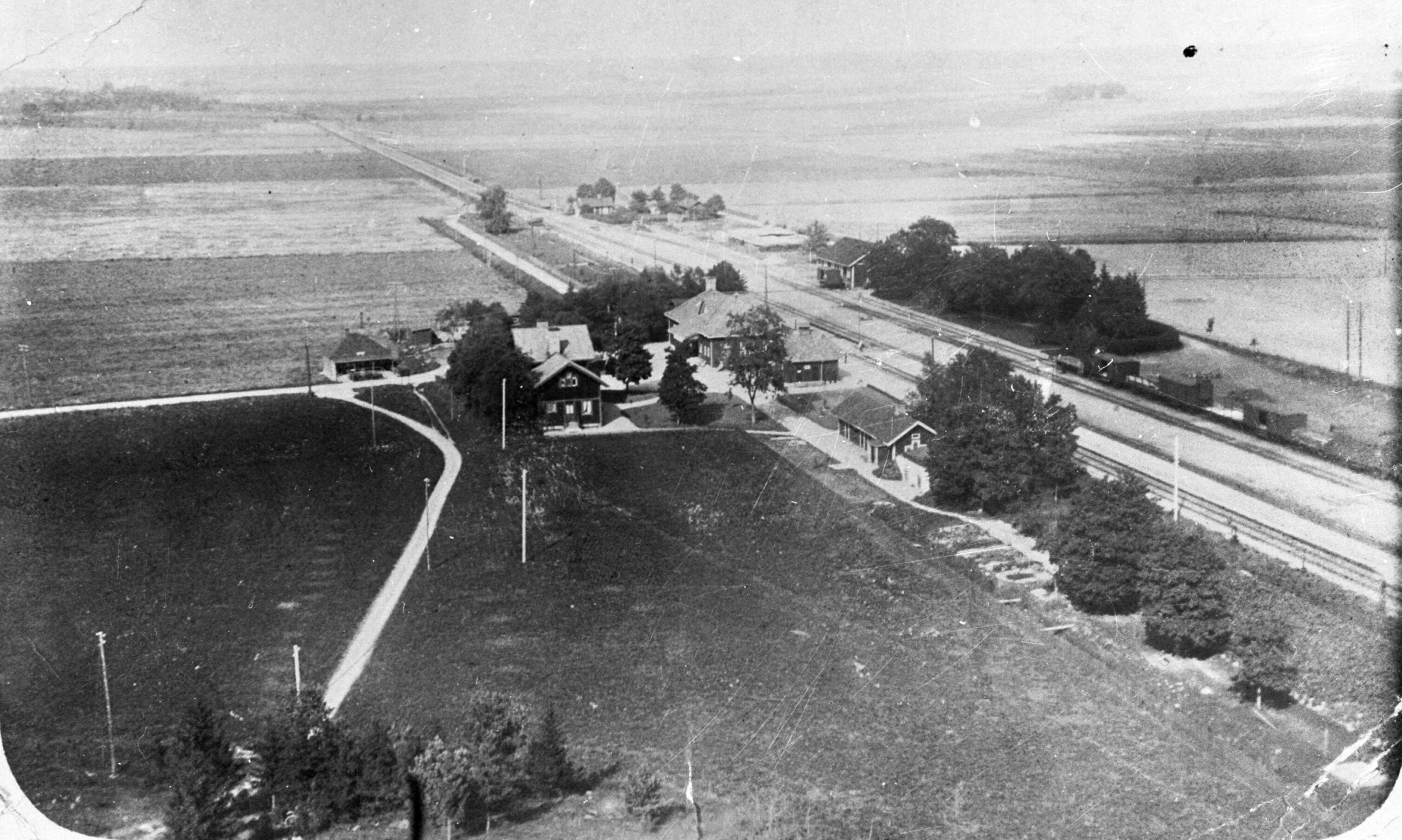 Svartvitt foto som visar järnvägen med stationshus och en handfull andra byggnader nära spåret. Resten är åker och äng.