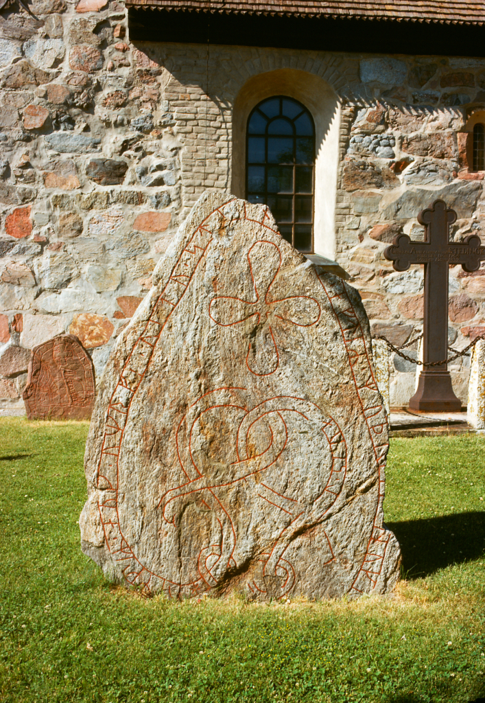 Runsten i förgrunden med ett upprest kors i bakgrunden, båda är placerade framför Lovö kyrkas fasad.