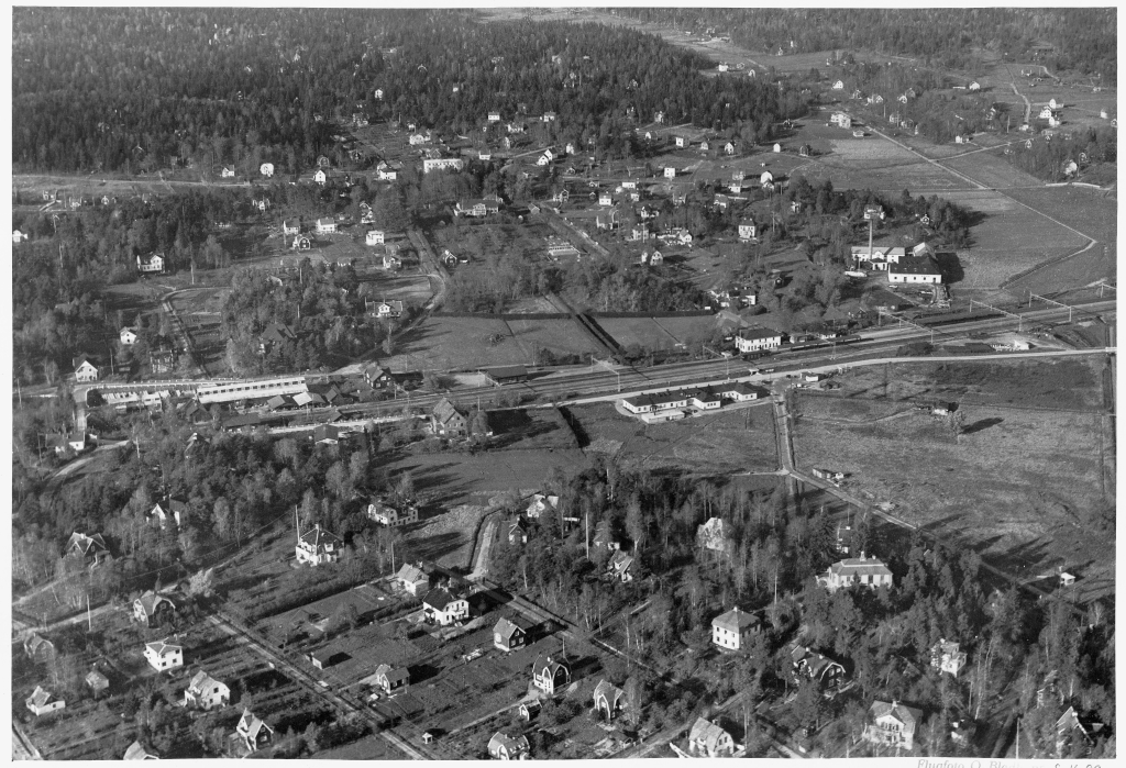 Ett flygfoto med villor, järnväg, stationshus och en fabriksbyggnad.