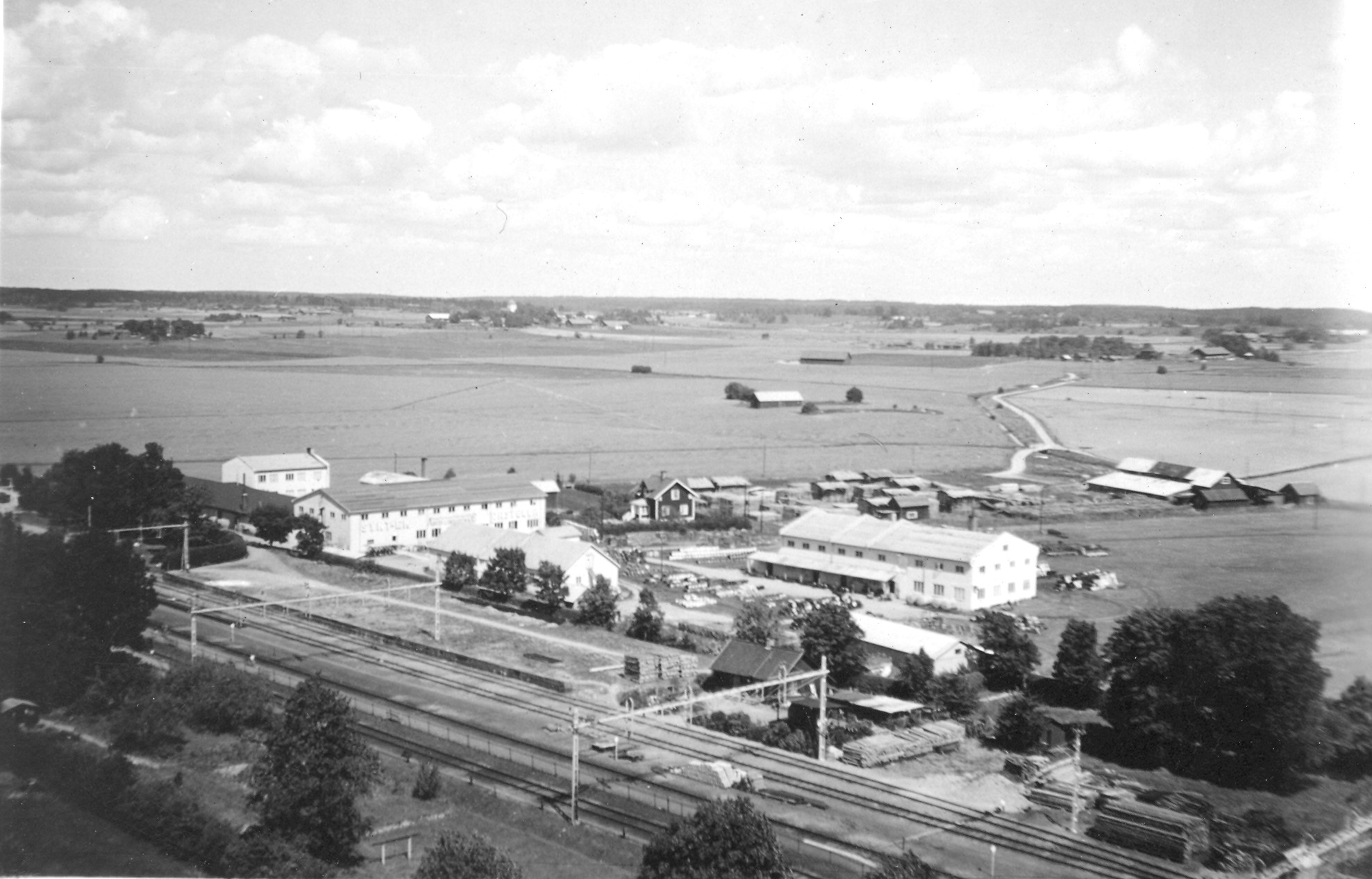 Ett svartvitt foto från hög höjd som visar tågspåren och några industribyggnader.