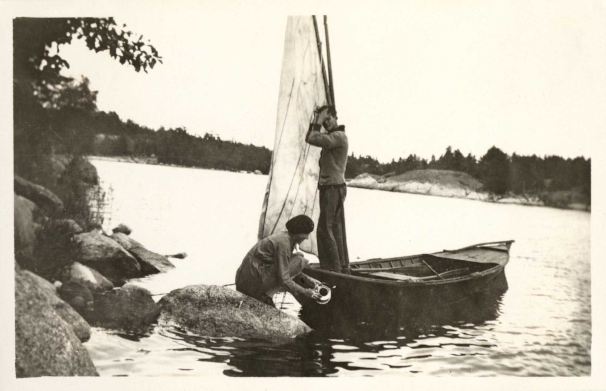 Svartvit bild på en person som står upp i en träbåt med segel, en person sitter hukad bredvid på en sten i vattnet