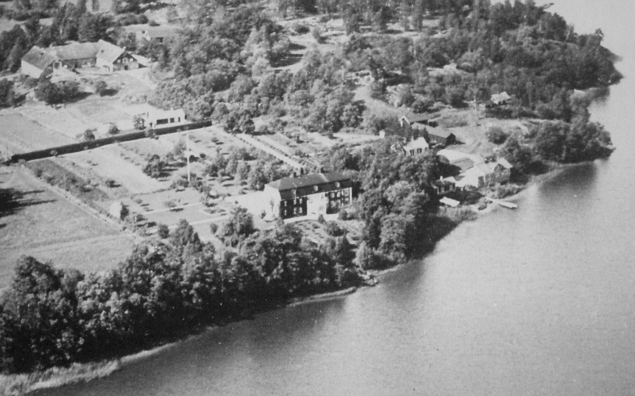 Svartvitt flygfoto där en herrgård med flera olika byggnader syns nära sjöstranden.