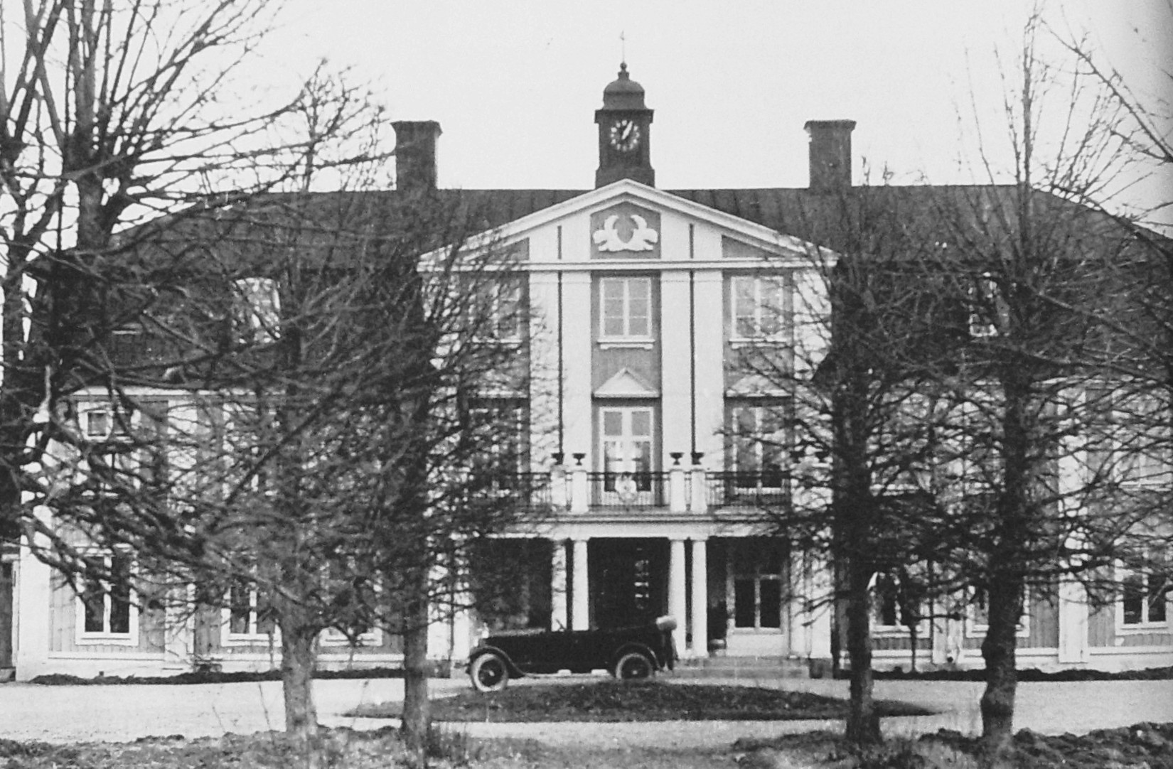 En svartvit bild av en herrgård med en gammal bil framför.