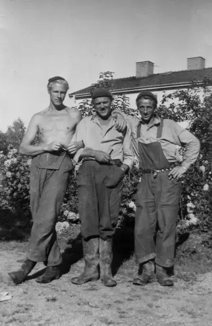 Tre män står uppställda framför kameran. En har bar överkropp, en annan bär hängselbyxor, alla har stövlar.