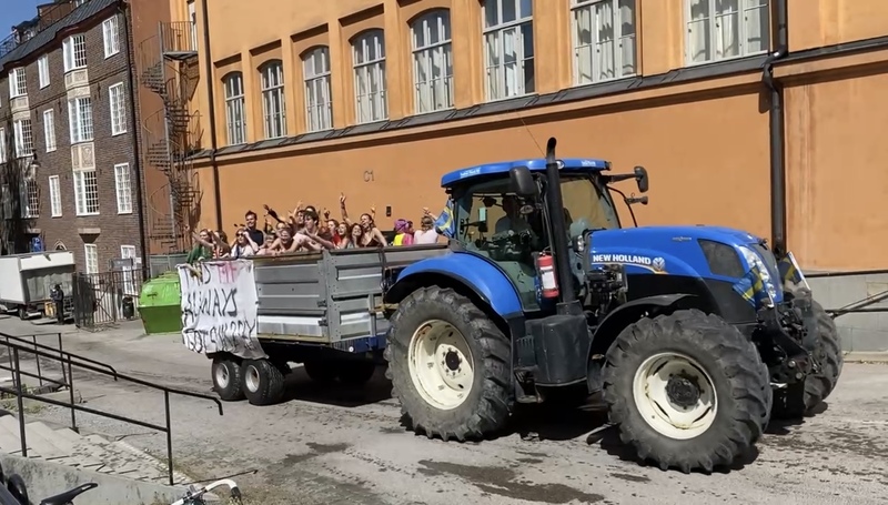 En blå traktor drar på en stor vagn, i vagnen står en grupp studentfirare.