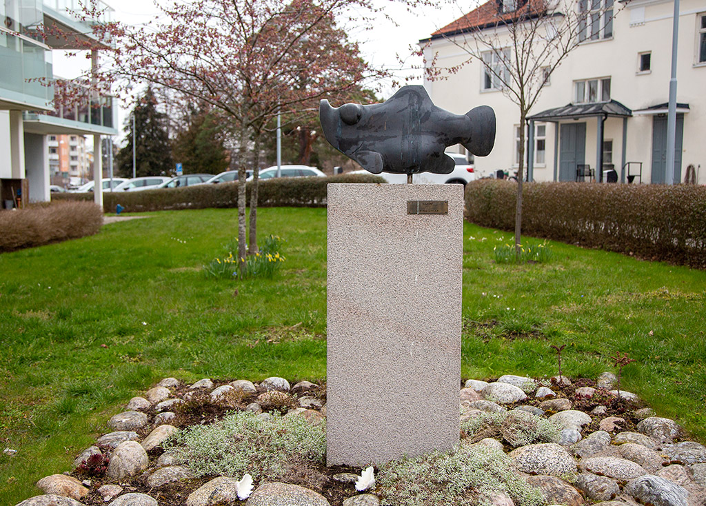 Skulptur i mörk brons av en fisk med trubbig framdel på en hög sten-sockel.