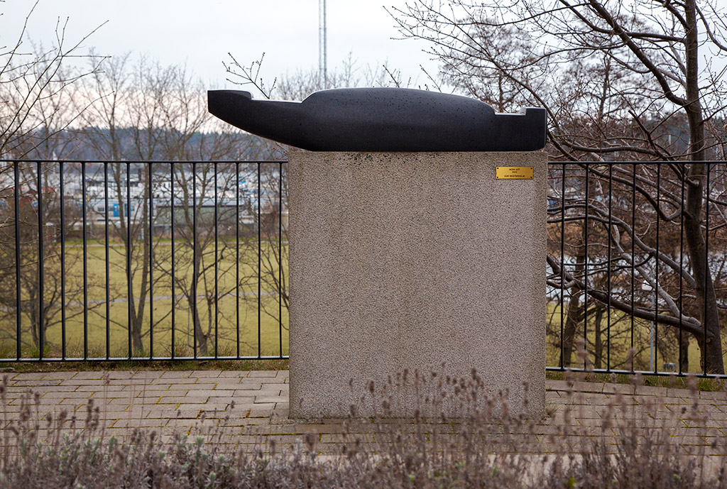 En svart abstrakt skulptur med avlång form står på en ljusgrå stensockel. I bakgrunden ett svart staket och utsikt mot hav och båtar.