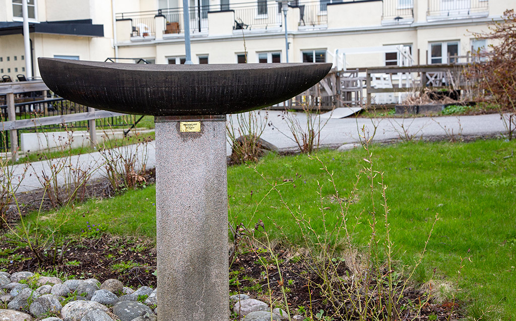 En mörk skulptur av en båt som står på en grå sockel av granit. Runtomkring finns grönt gräs och spirande rosenbuskar. I bakgrunden syns hus med lägenheter.