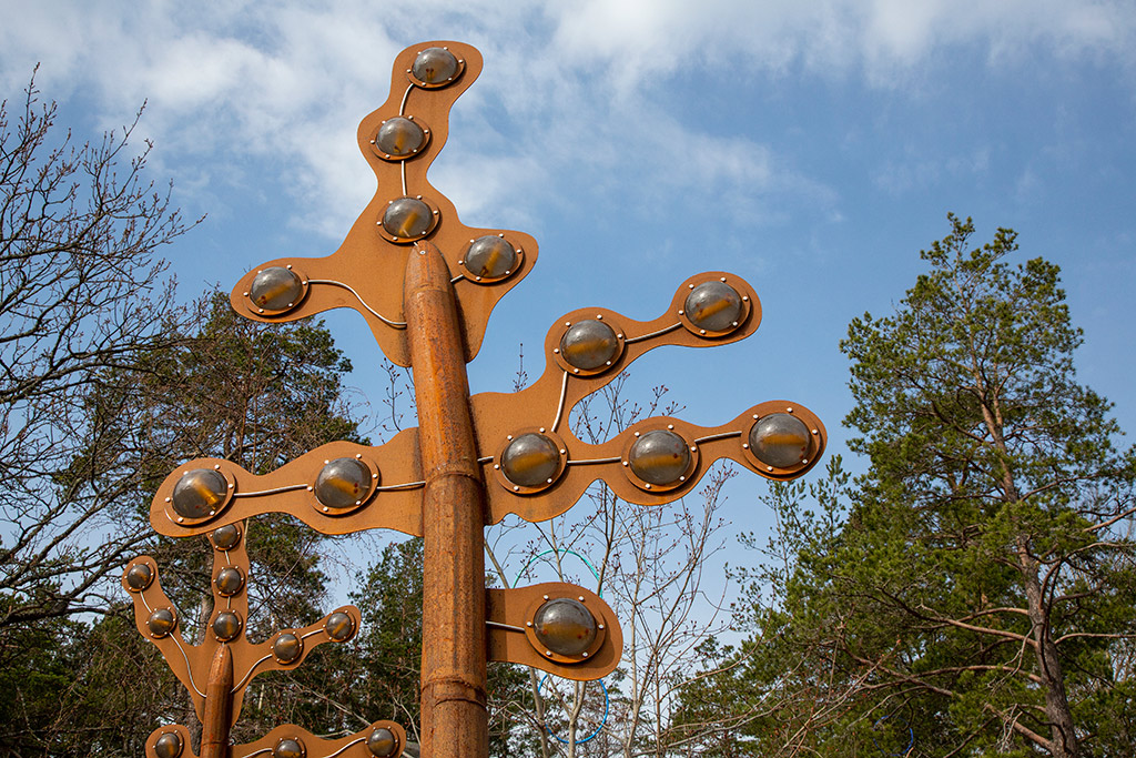 Brun skulptur som liknar tång mot en blå himmel med trädtoppar runtomkring.