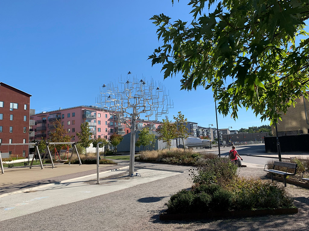 Ett träd av stål som står i en park omgiven av gröna träd och hus-