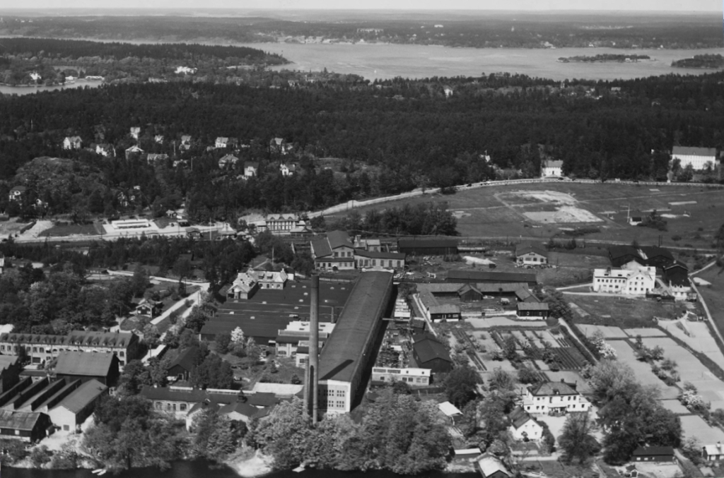 Ett svartvitt flygfoto där en lång industribyggnad med hög skorsten dominerar bilden.