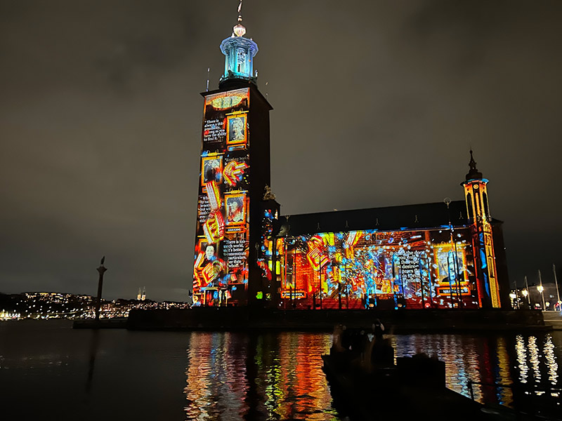 Fotografi av Stadshuset med ljusinstallation med porträtt av kvinnor i starka färger, mest orange.