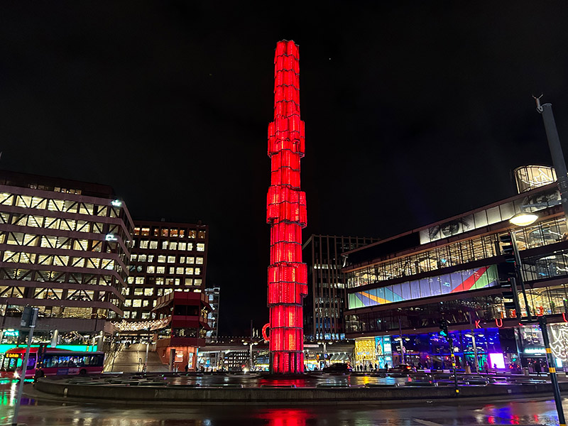 Röd, lysande, hög skulptur i en storstad om kvällen.