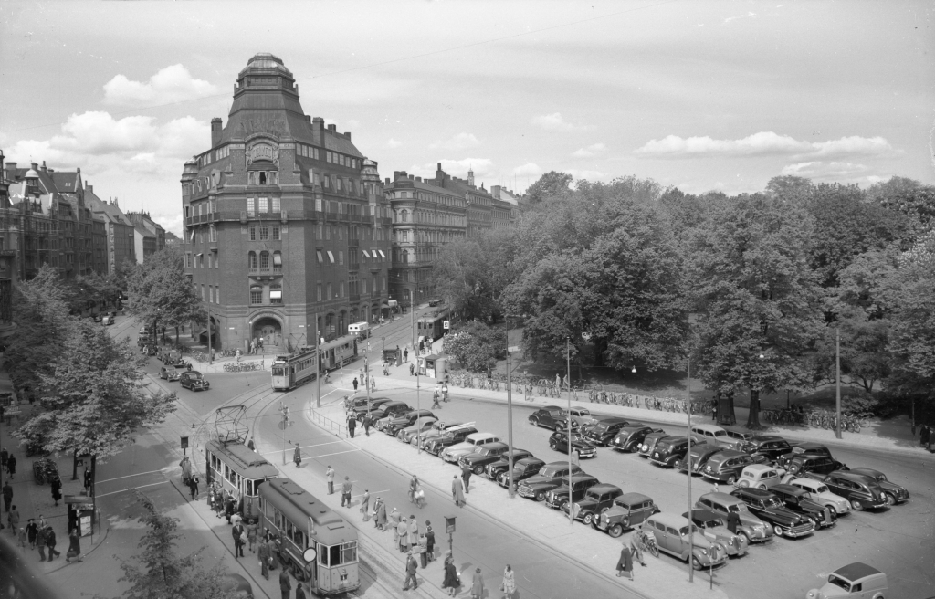 Svartvitt foto med stadsmiljö med ålderdomliga bilar och två spårvagnar.