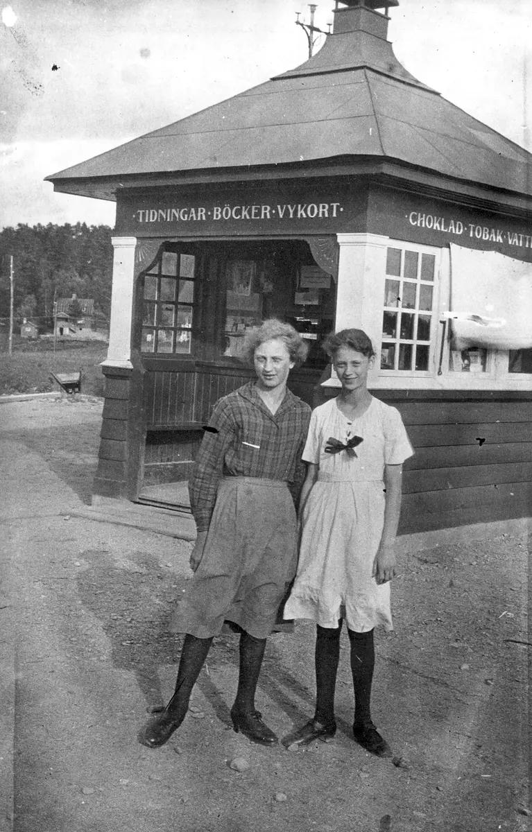 Två flickor står uppställda framför en kiosk med pyramidformat tak på detta svartvita foto. På kiosken står skrivet: Tidningar, böcker, vykort, choklad, Tobak, Vatt....