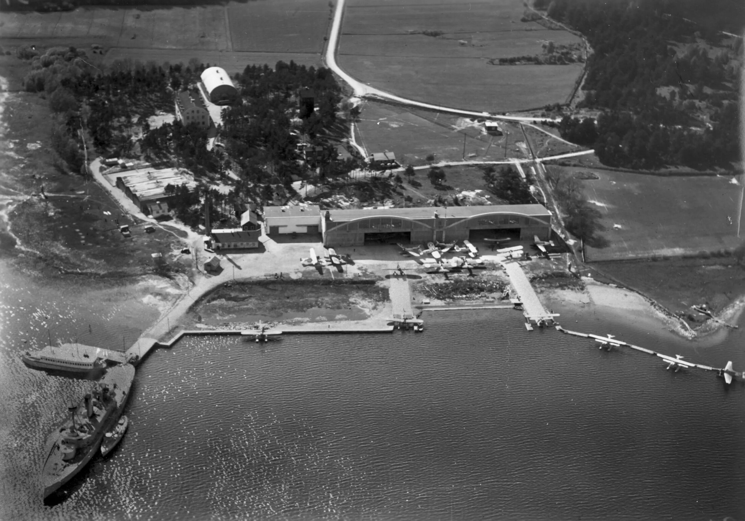 Svartvitt flygfoto med flygplan och båtar på vattnet, hangarer och andra byggnader på land.