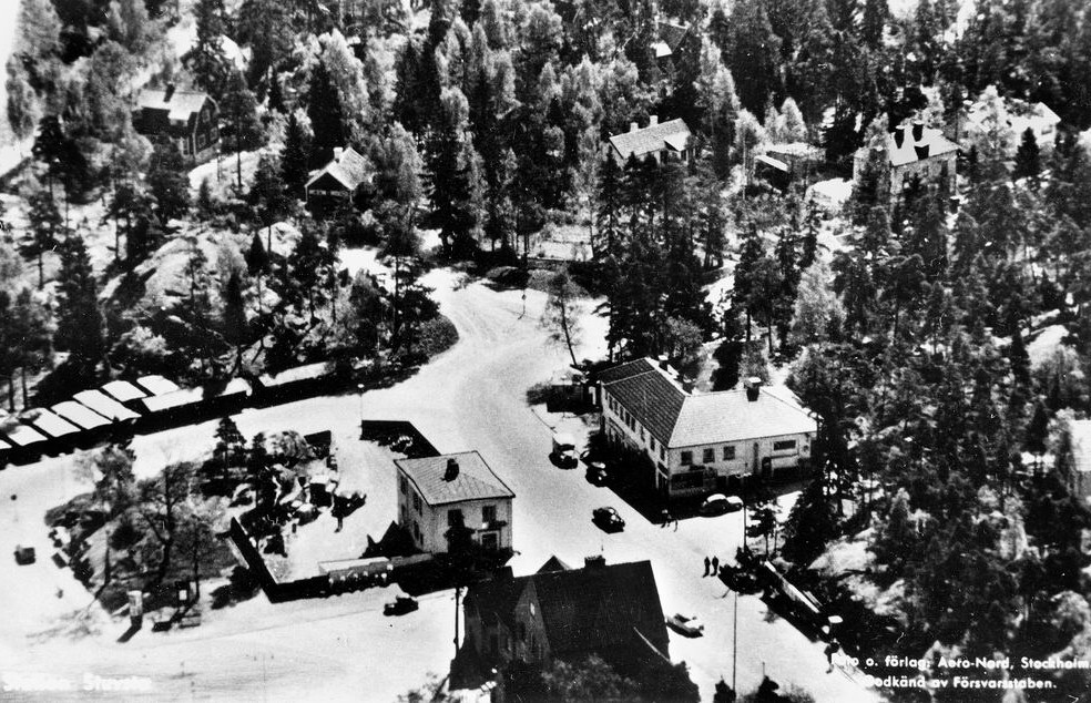 En svartvit flygbild med små låga hus och många träd.