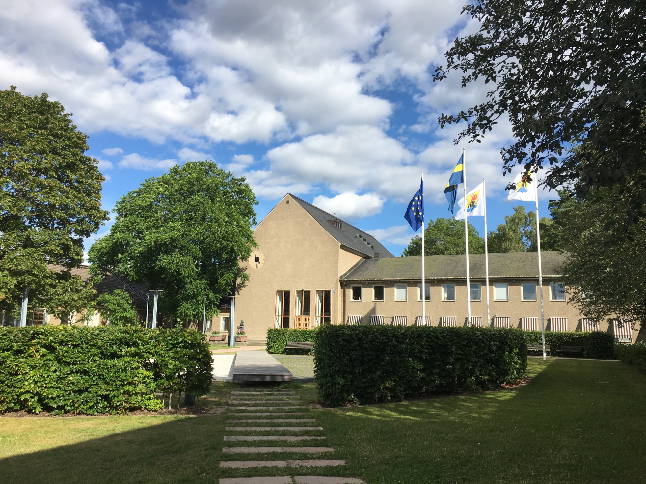 På bilden syns kommunhuset med en hög gavel och längre byggnad till höger. Kommunens vapen, EU-flaggan och Sverigeflaggan vajar på var sin stång.