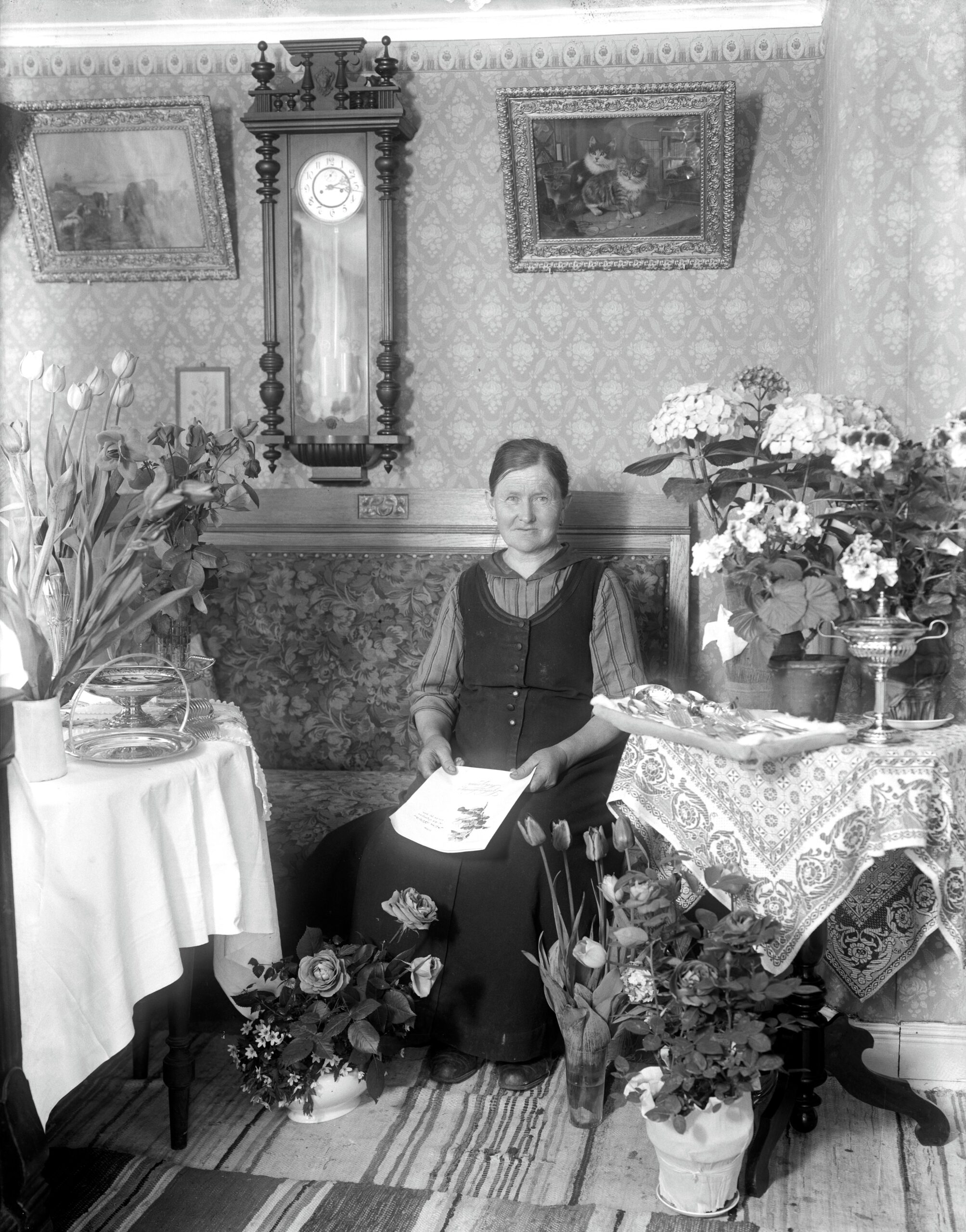 Svartvitt foto där Mina sitter i en salong omgiven av blombuketter. Trasmattor på golvet, medaljongtapet, väggur och tavlor på väggen.